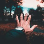 A person's hand extends towards a blurred, colorful outdoor autumn scene, wearing a wristwatch.